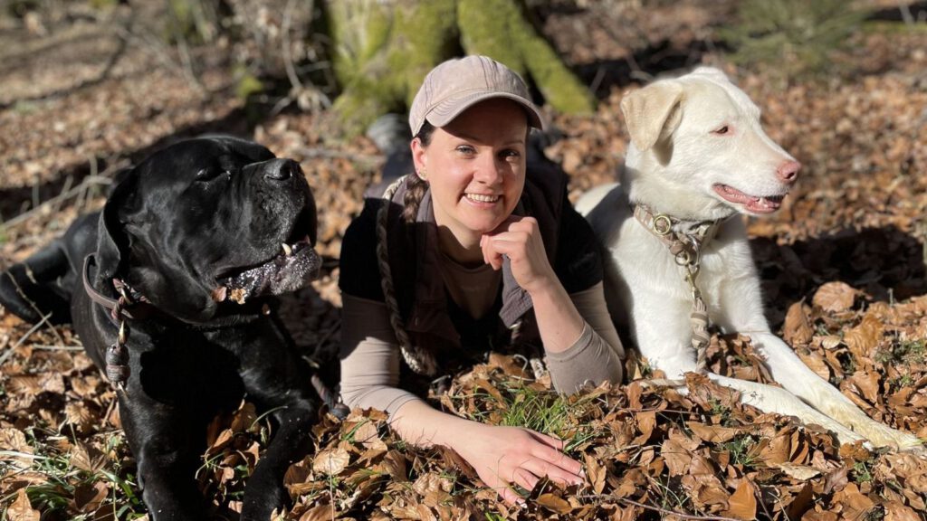 Foto: Frau liegt fröhlich mit ihren Hunden auf dem Waldboden.